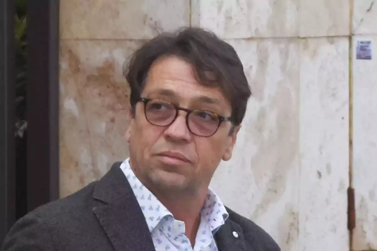 Hombre de cabello oscuro y gafas con expresión seria, vestido con saco gris y camisa clara, de pie frente a una pared de mármol. Hombre de cabello oscuro y gafas con expresión seria, vestido con saco gris y camisa clara, de pie frente a una pared de mármol.