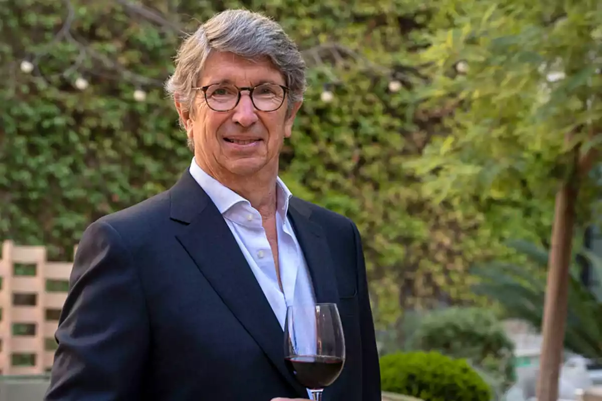 Hombre de cabello canoso y gafas sosteniendo una copa de vino tinto en un jardín Hombre de cabello canoso y gafas sosteniendo una copa de vino tinto en un jardín