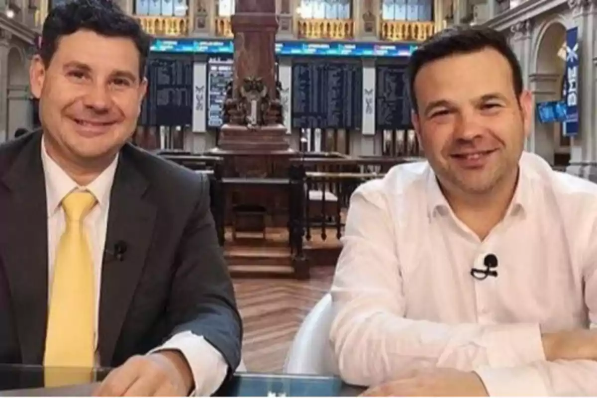 Dos hombres sonrientes sentados en una mesa dentro de un edificio elegante con pantallas de información financiera al fondo Dos hombres sonrientes sentados en una mesa dentro de un edificio elegante con pantallas de información financiera al fondo