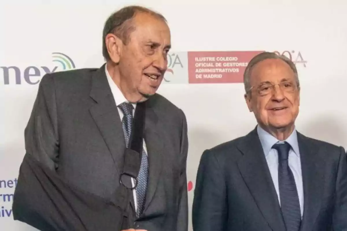 Dos hombres mayores con traje sonríen frente a un fondo con logotipos uno de ellos lleva el brazo en cabestrillo negro Dos hombres mayores con traje sonríen frente a un fondo con logotipos uno de ellos lleva el brazo en cabestrillo negro