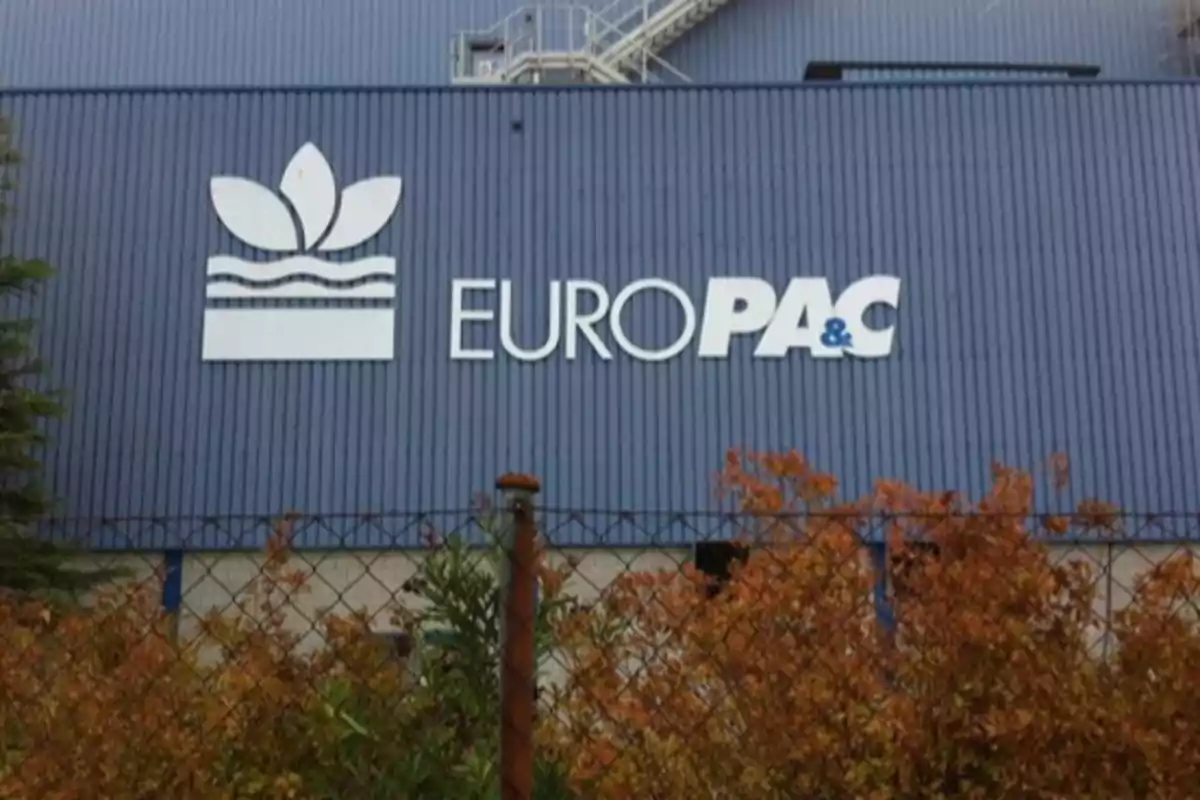 Fachada de edificio industrial azul con el logo y el nombre EURO PAC acompañado de arbustos y una cerca en primer plano Fachada de edificio industrial azul con el logo y el nombre EURO PAC acompañado de arbustos y una cerca en primer plano