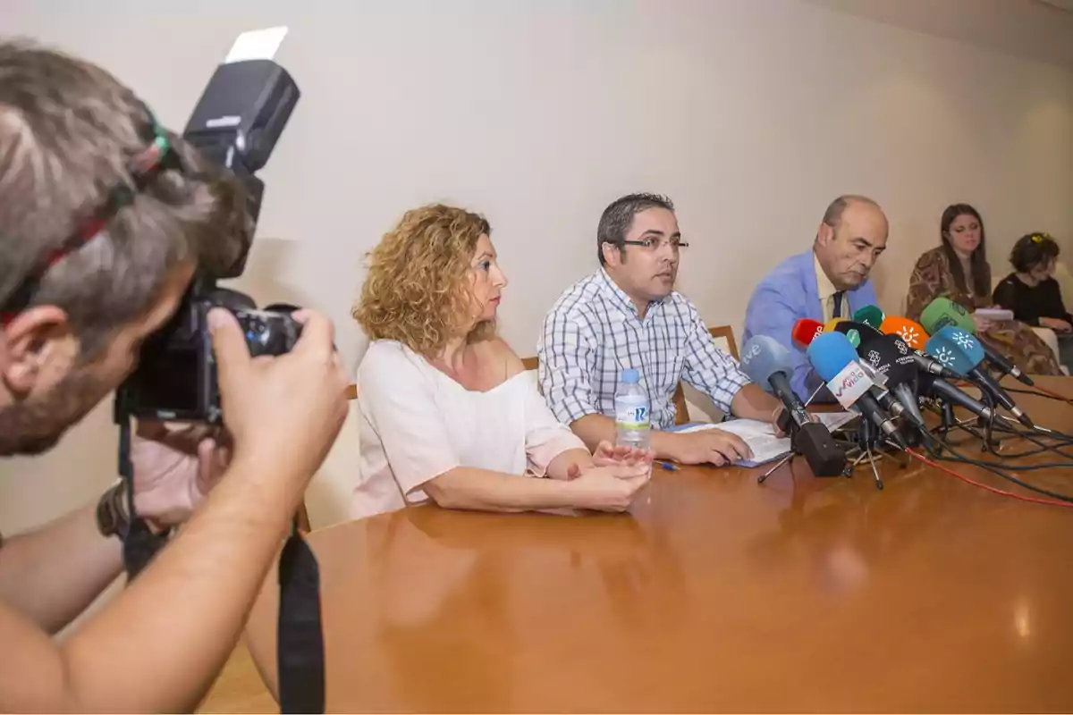 Un grupo de personas sentadas en una mesa durante una rueda de prensa con varios micrófonos y un fotógrafo capturando el momento. Un grupo de personas sentadas en una mesa durante una rueda de prensa con varios micrófonos y un fotógrafo capturando el momento.