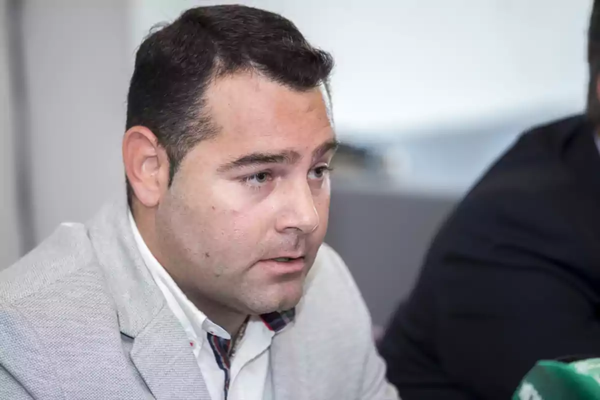 Hombre de cabello corto y oscuro vistiendo saco gris claro y camisa blanca sentado en un entorno interior Hombre de cabello corto y oscuro vistiendo saco gris claro y camisa blanca sentado en un entorno interior