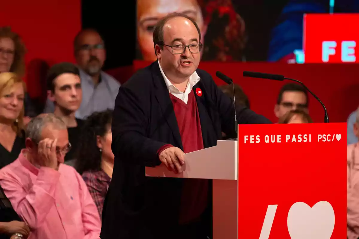 Hombre con gafas y chaqueta oscura hablando en un atril rojo con el lema Fes Que Passi PSC, rodeado de personas en un evento político. Hombre con gafas y chaqueta oscura hablando en un atril rojo con el lema Fes Que Passi PSC, rodeado de personas en un evento político.