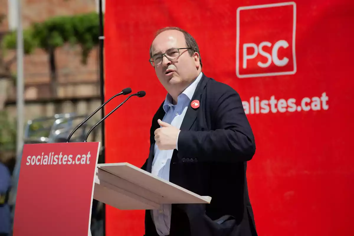 Hombre con gafas hablando en un mitin político del PSC frente a un atril rojo con el logo y la web del partido Hombre con gafas hablando en un mitin político del PSC frente a un atril rojo con el logo y la web del partido
