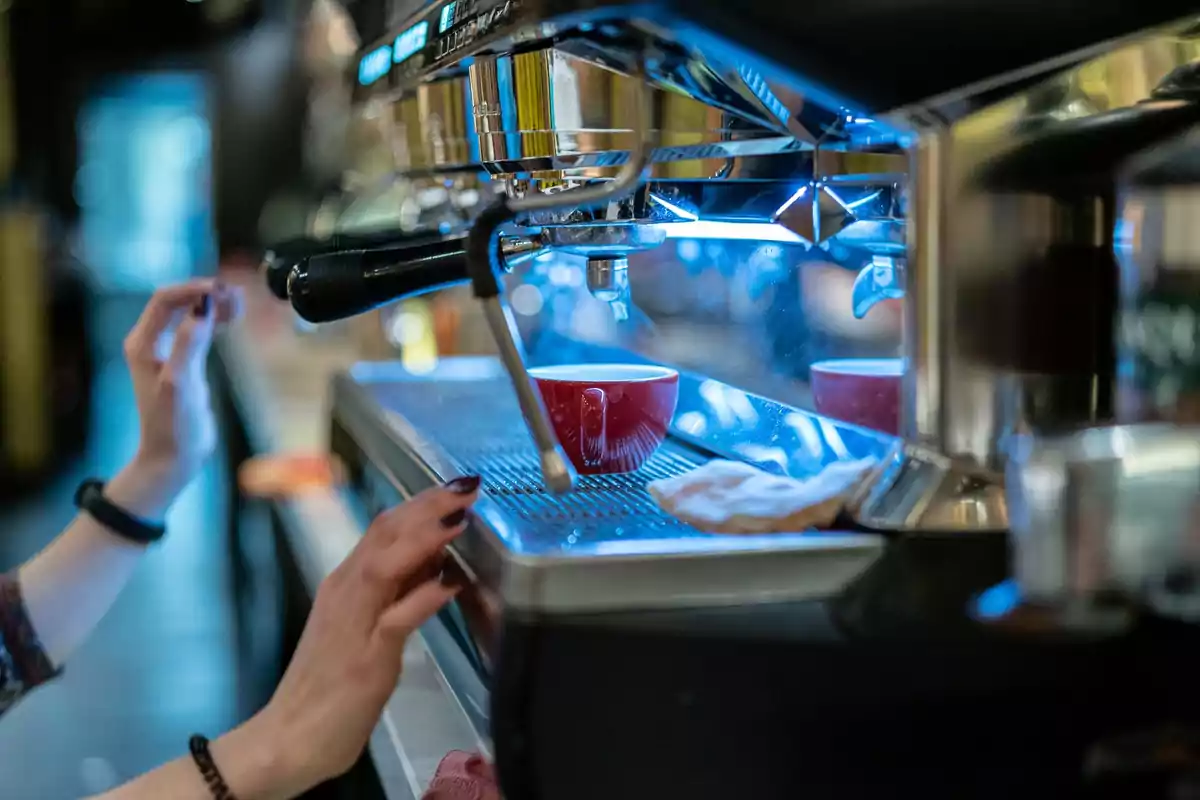 Persona preparando café con una máquina profesional mientras una taza roja espera debajo del dispensador Persona preparando café con una máquina profesional mientras una taza roja espera debajo del dispensador