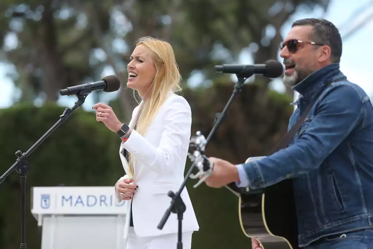 Una mujer vestida de blanco canta frente a un micrófono mientras un hombre con chaqueta de mezclilla toca la guitarra y también canta al aire libre Una mujer vestida de blanco canta frente a un micrófono mientras un hombre con chaqueta de mezclilla toca la guitarra y también canta al aire libre