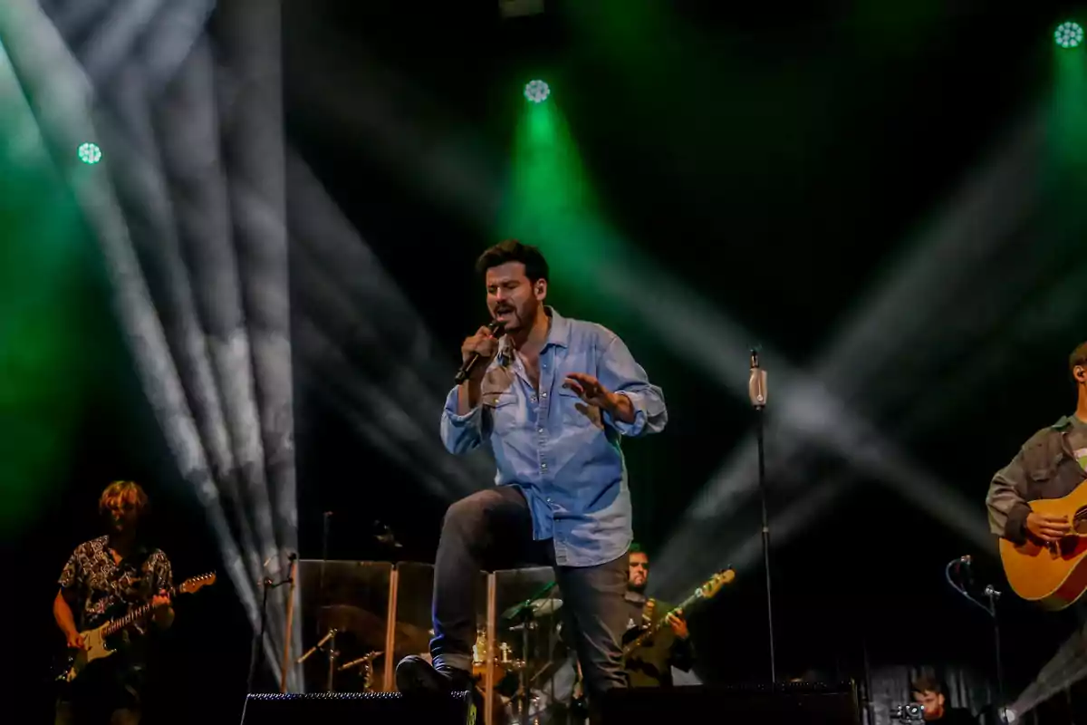 Cantante interpretando una canción en el escenario acompañado de su banda con luces verdes y blancas de fondo Cantante interpretando una canción en el escenario acompañado de su banda con luces verdes y blancas de fondo