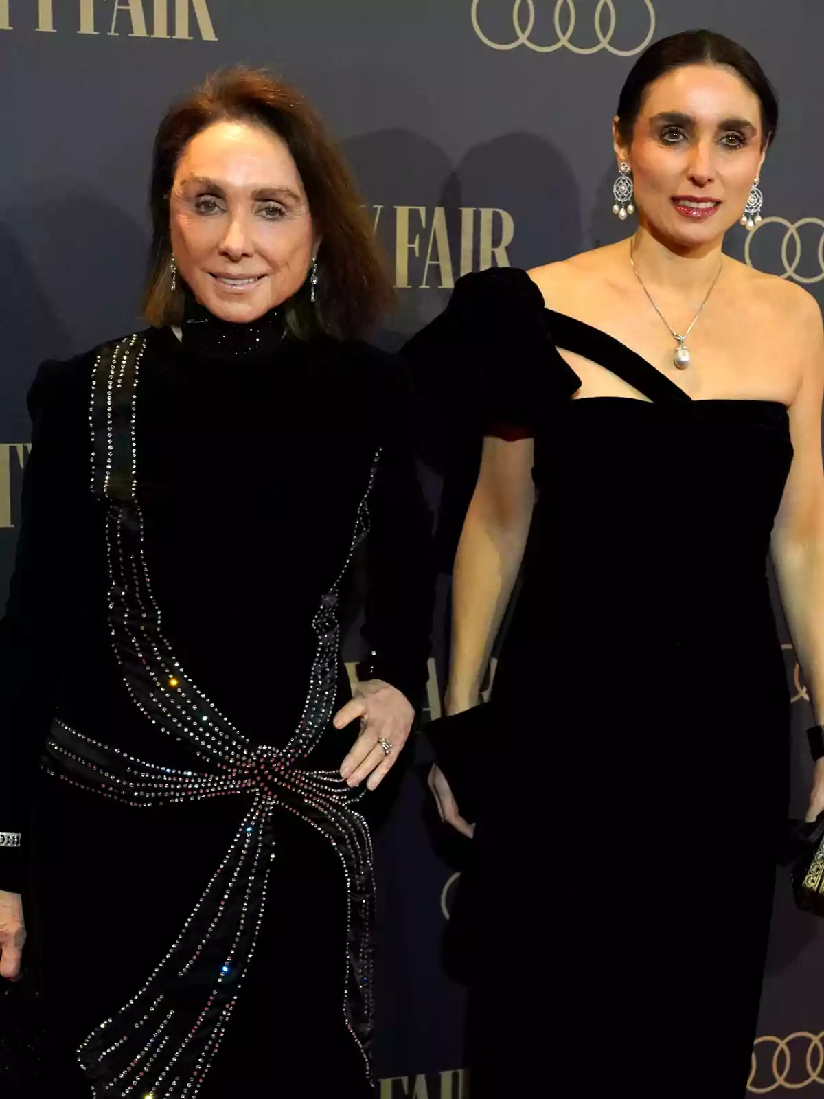 Dos mujeres posan en un evento elegante con vestidos negros y joyería llamativa frente a un fondo con el logo de Vanity Fair y Audi Dos mujeres posan en un evento elegante con vestidos negros y joyería llamativa frente a un fondo con el logo de Vanity Fair y Audi