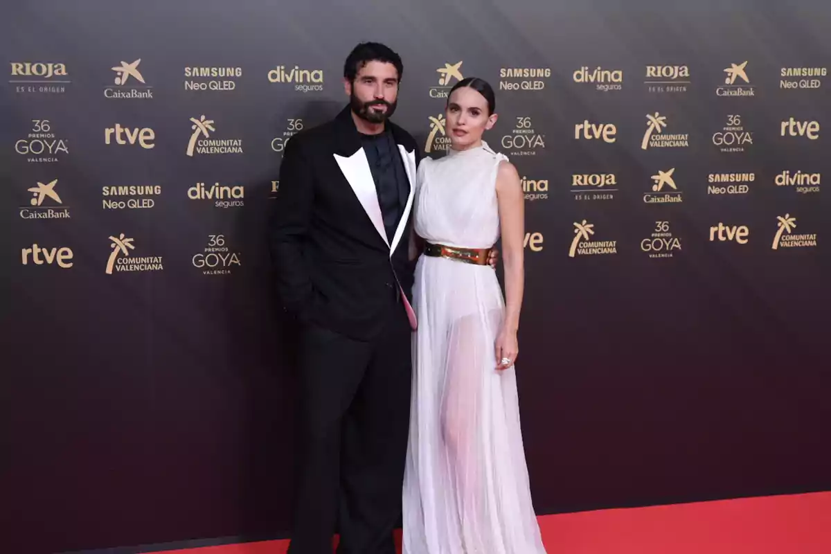 Una pareja posa elegantemente en la alfombra roja de los Premios Goya frente a un fondo con logotipos dorados de patrocinadores y organizadores del evento Una pareja posa elegantemente en la alfombra roja de los Premios Goya frente a un fondo con logotipos dorados de patrocinadores y organizadores del evento