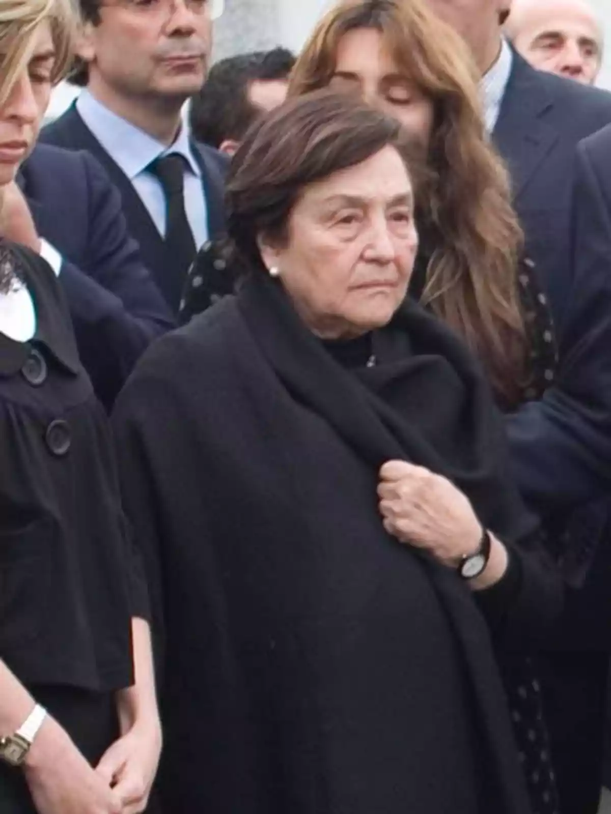 Mujer mayor vestida de negro rodeada de personas en lo que parece ser un evento solemne Mujer mayor vestida de negro rodeada de personas en lo que parece ser un evento solemne