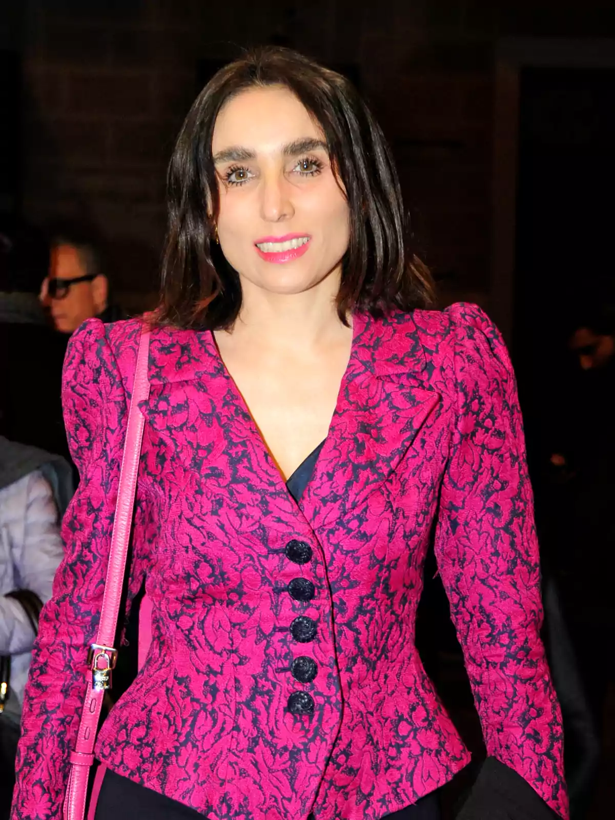 Mujer de cabello castaño oscuro y lacio, sonriente, usando un saco rosa con estampado floral y botones negros, lleva un bolso rosa al hombro y está en un ambiente interior con personas al fondo. Mujer de cabello castaño oscuro y lacio, sonriente, usando un saco rosa con estampado floral y botones negros, lleva un bolso rosa al hombro y está en un ambiente interior con personas al fondo.