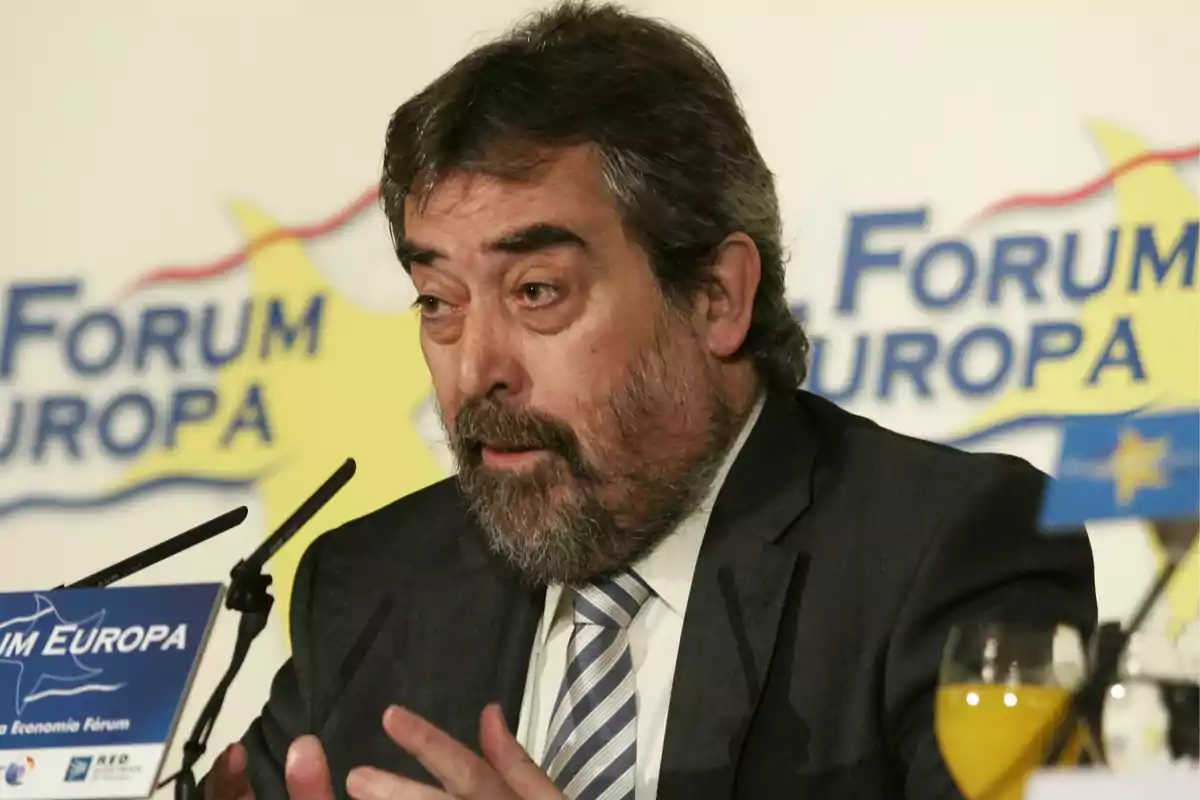 Hombre de cabello y barba canosos hablando en un evento llamado Forum Europa con un micrófono y un vaso de jugo en la mesa Hombre de cabello y barba canosos hablando en un evento llamado Forum Europa con un micrófono y un vaso de jugo en la mesa