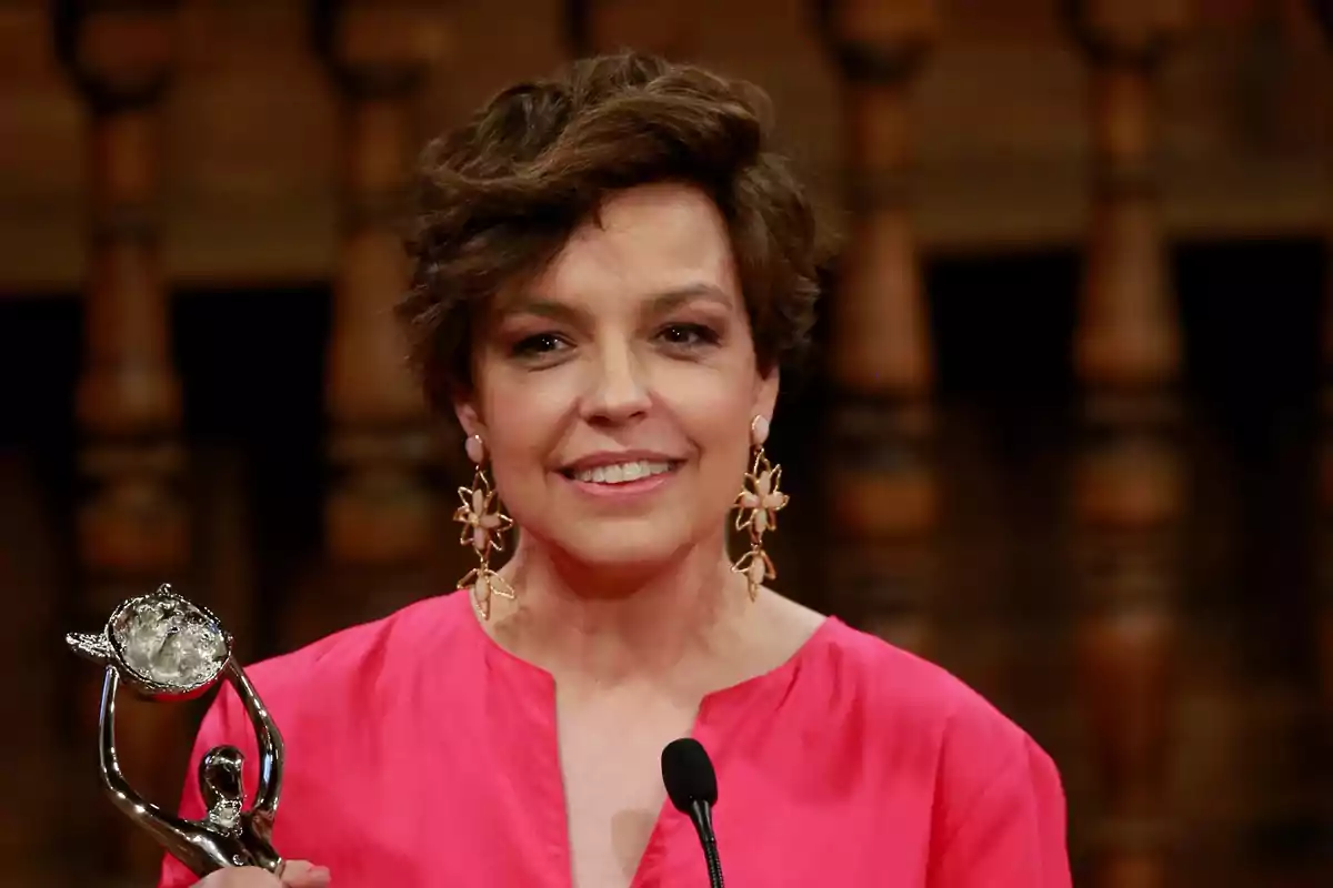 Mujer con cabello corto y vestido rosa sosteniendo un trofeo y sonriendo frente a un micrófono