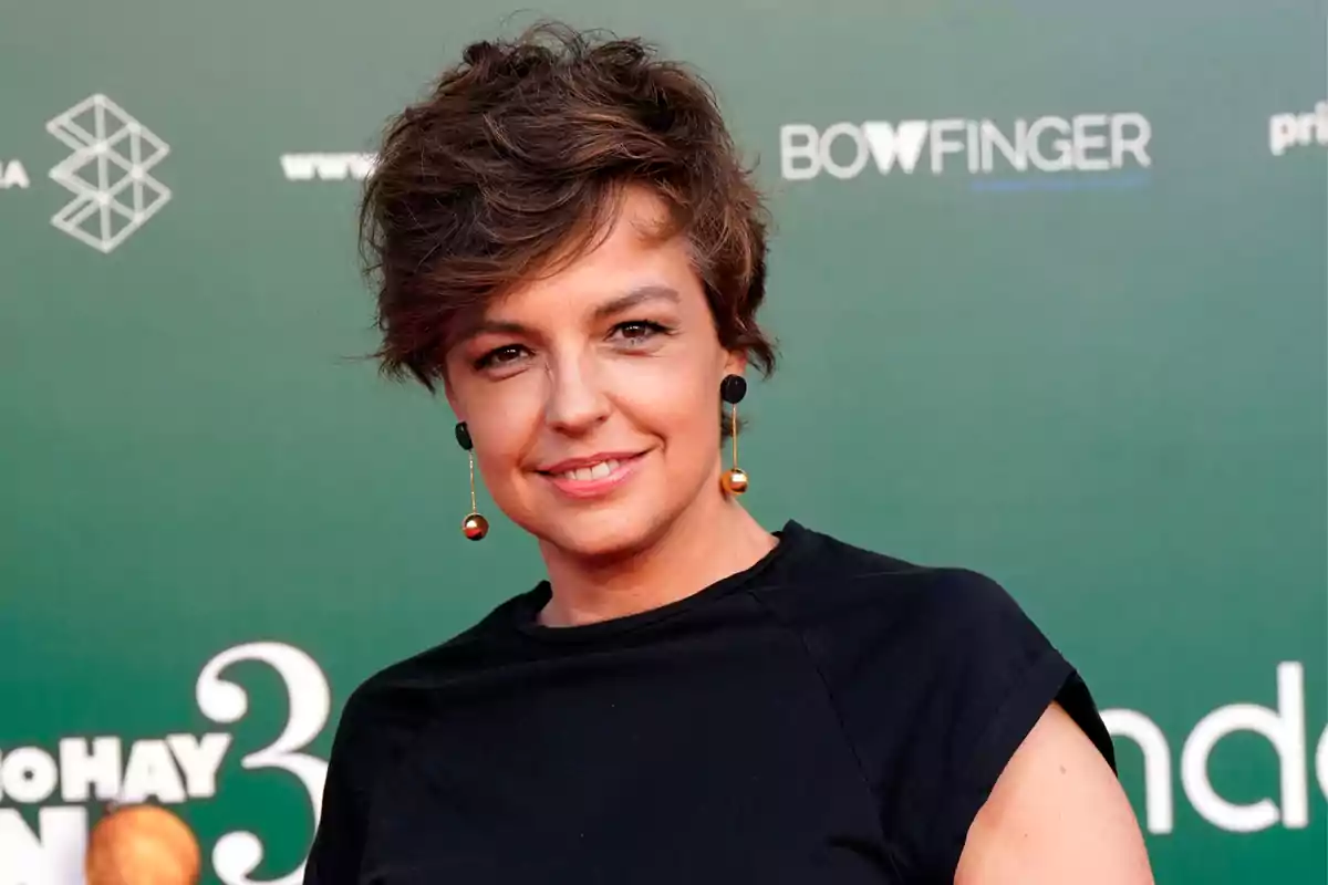 Mujer de cabello corto y castaño con pendientes largos y camiseta negra posando frente a un fondo verde en un evento