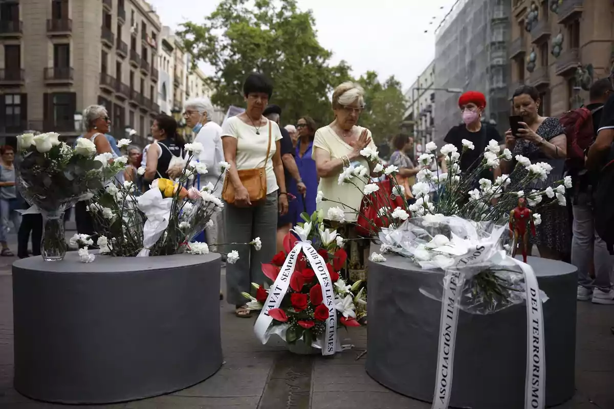 Personas reunidas en una calle alrededor de arreglos florales en un acto de homenaje o conmemoración
