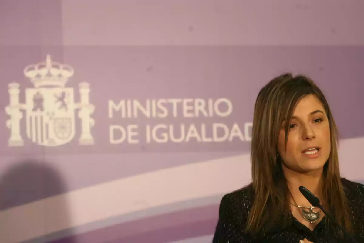 Mujer hablando frente a un micrófono con un fondo que muestra el escudo de España y el texto Ministerio de Igualdad
