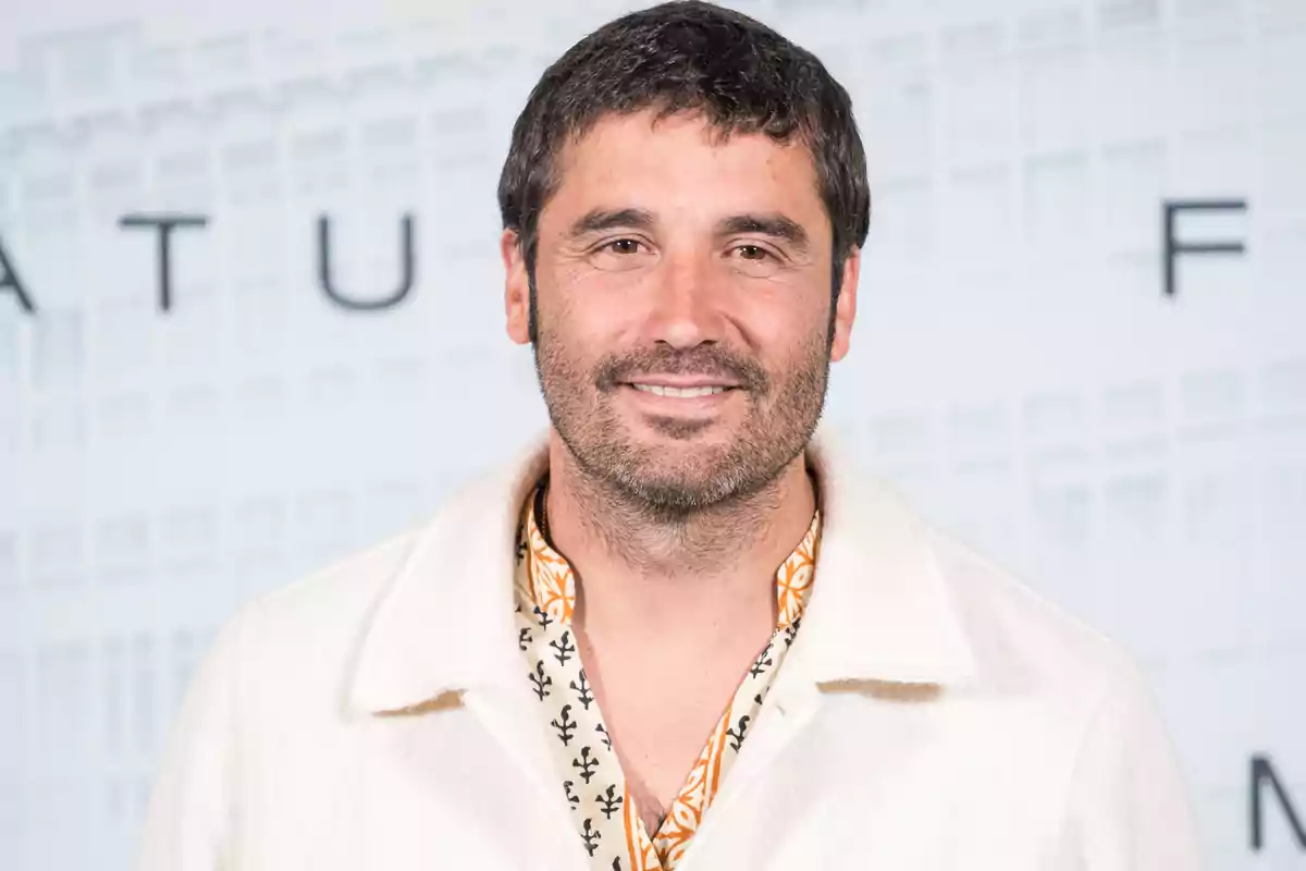 Hombre de cabello corto y barba con chaqueta blanca y camisa estampada sonríe frente a un fondo claro desenfocado Hombre de cabello corto y barba con chaqueta blanca y camisa estampada sonríe frente a un fondo claro desenfocado