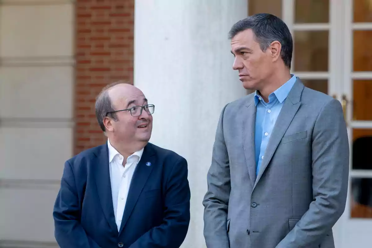 Dos hombres con traje conversan de pie frente a un edificio con columnas y una puerta de vidrio al fondo Dos hombres con traje conversan de pie frente a un edificio con columnas y una puerta de vidrio al fondo