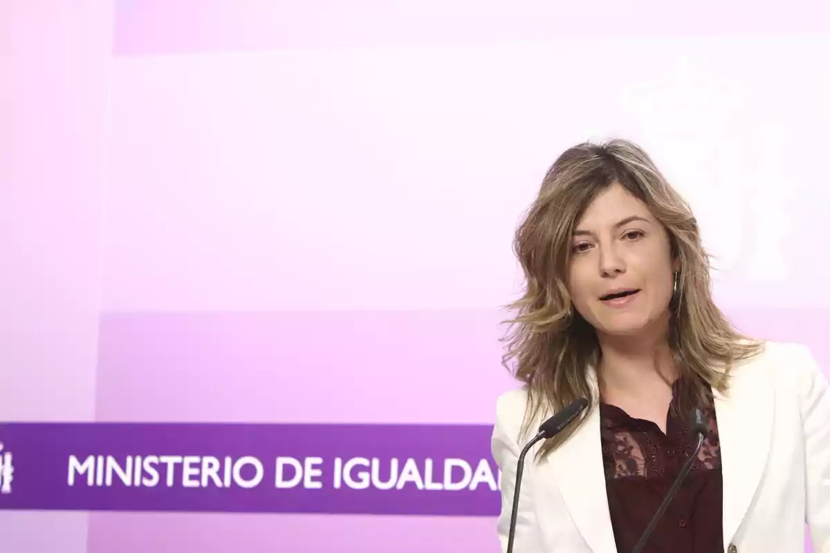 Mujer hablando frente a un micrófono con fondo del Ministerio de Igualdad en tonos morados Mujer hablando frente a un micrófono con fondo del Ministerio de Igualdad en tonos morados