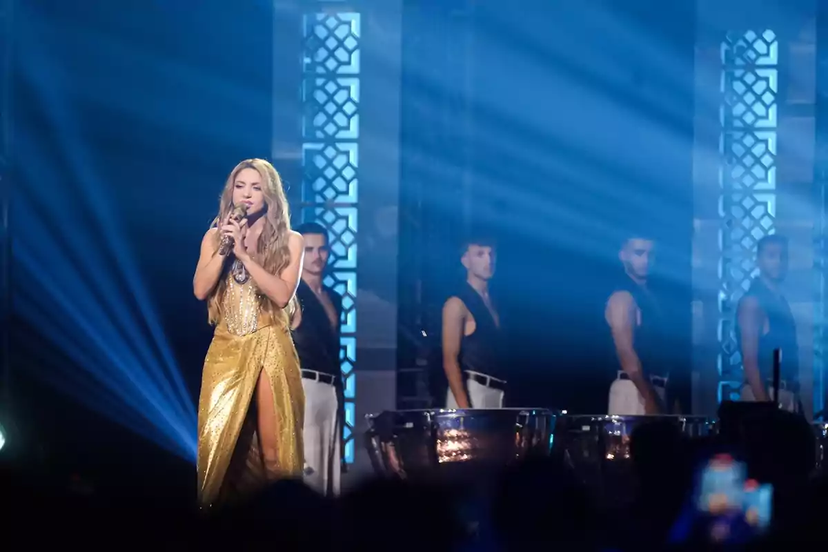 Cantante con vestido dorado interpretando una canción en el escenario acompañada de músicos y luces azules de fondo Cantante con vestido dorado interpretando una canción en el escenario acompañada de músicos y luces azules de fondo