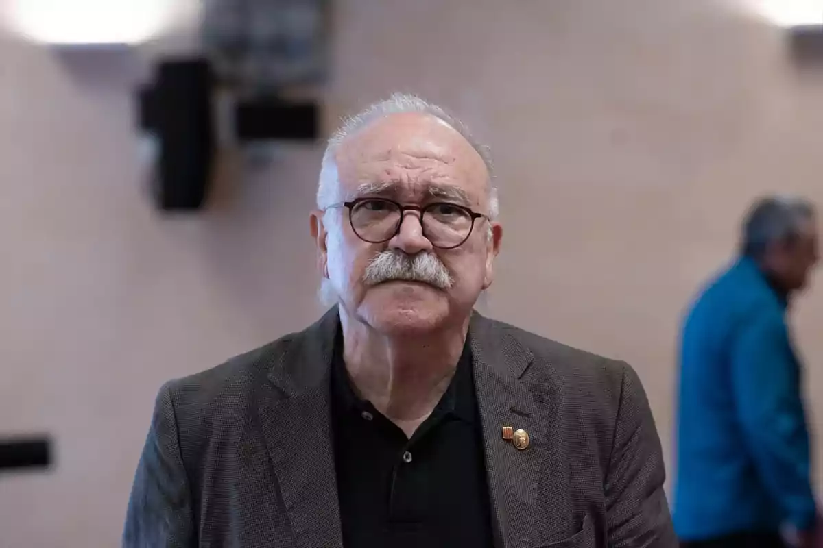 Hombre mayor con gafas y bigote canoso viste saco gris y camisa negra en un ambiente interior desenfocado