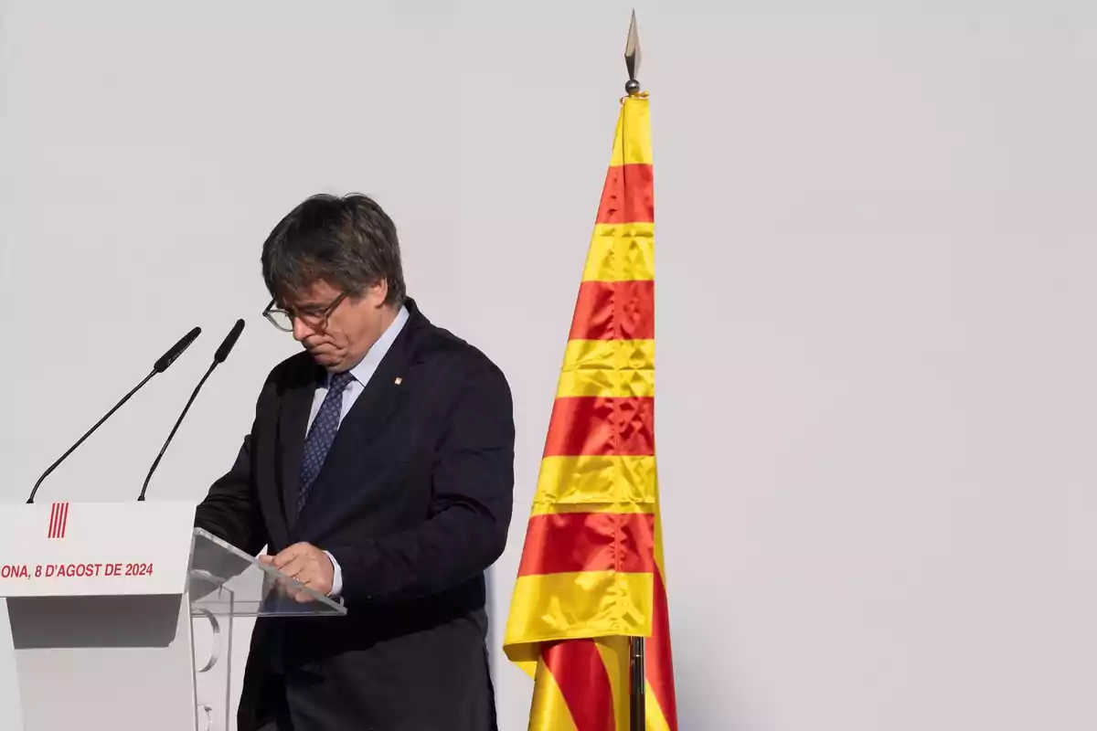 Hombre con gafas y traje oscuro leyendo un discurso en un atril junto a una bandera amarilla con franjas rojas