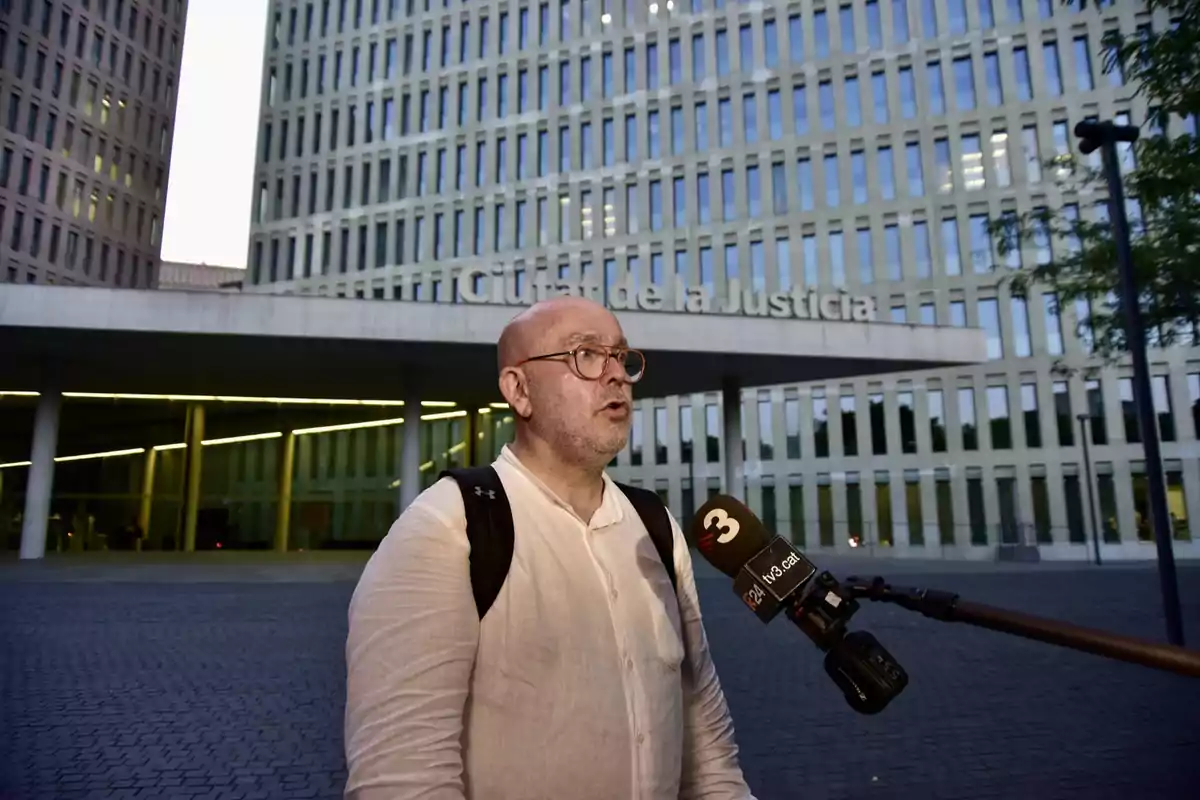 Hombre con gafas y camisa clara hablando frente a un micrófono de televisión en la entrada de un edificio moderno llamado Ciudad de la Justicia