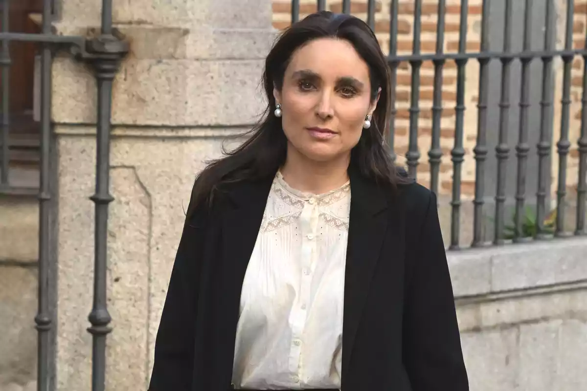 Mujer de cabello oscuro y suelto vestida con blusa blanca y saco negro de pie frente a una pared de piedra y una reja de metal Mujer de cabello oscuro y suelto vestida con blusa blanca y saco negro de pie frente a una pared de piedra y una reja de metal