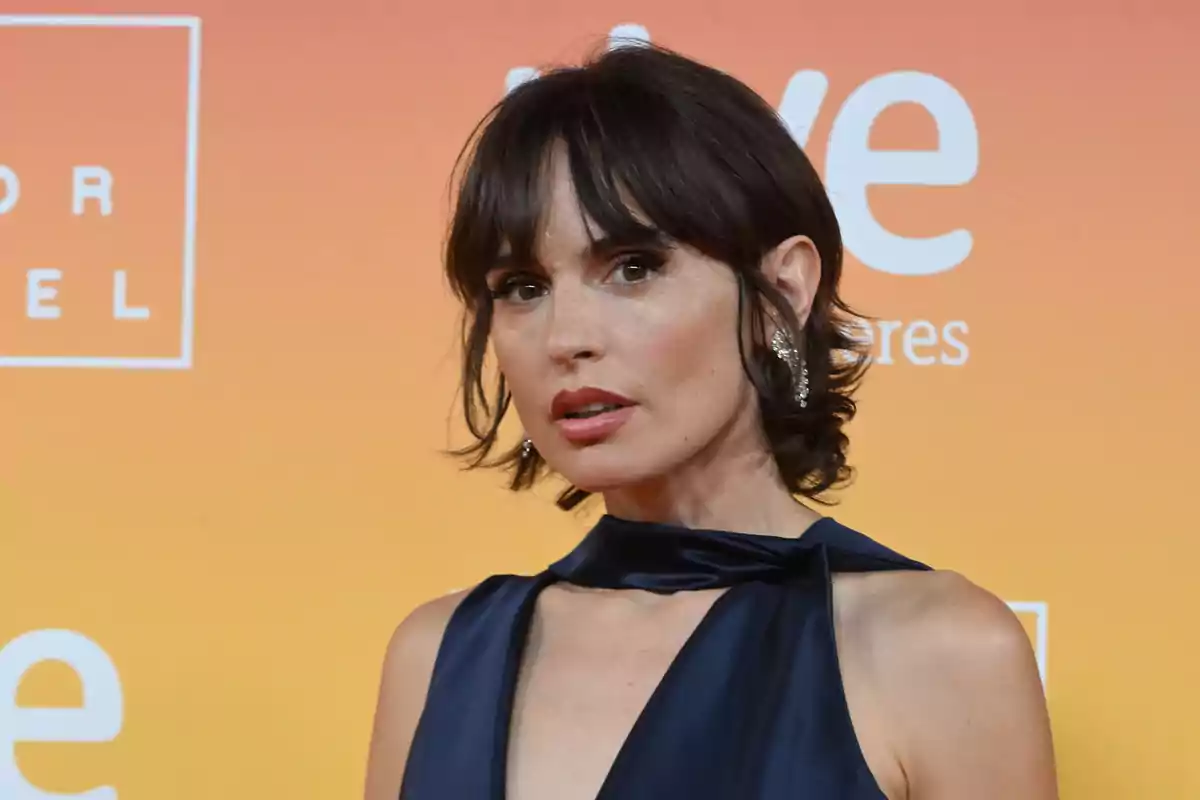 Mujer de cabello corto y oscuro con vestido azul elegante posando frente a un fondo naranja en un evento