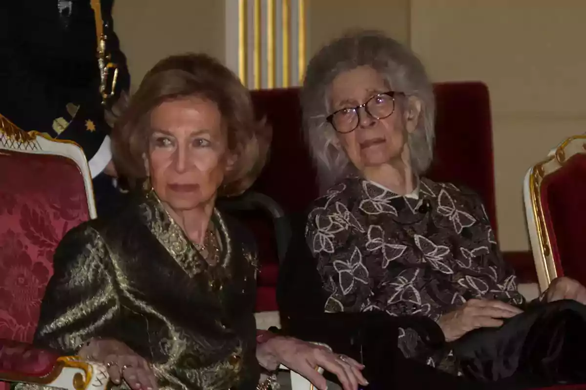 Two elegantly dressed older women are sitting in red armchairs in a formal setting. Two elegantly dressed older women are sitting in red armchairs in a formal setting.