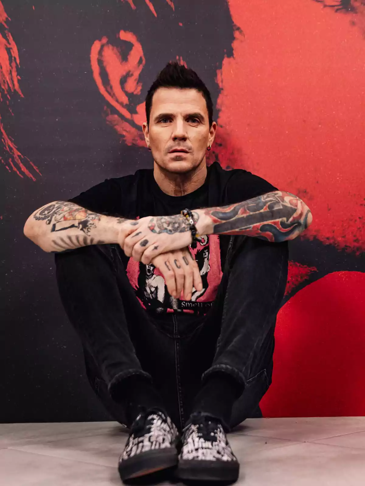 Hombre de cabello corto y oscuro con tatuajes en ambos brazos, sentado en el suelo con las piernas cruzadas, vistiendo camiseta negra y pantalón oscuro, frente a un fondo artístico en tonos rojo y negro Hombre de cabello corto y oscuro con tatuajes en ambos brazos, sentado en el suelo con las piernas cruzadas, vistiendo camiseta negra y pantalón oscuro, frente a un fondo artístico en tonos rojo y negro