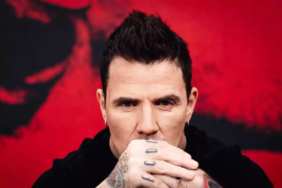 Hombre de cabello corto y oscuro con tatuajes en las manos y aretes, apoyando el rostro sobre sus manos frente a un fondo rojo difuminado Hombre de cabello corto y oscuro con tatuajes en las manos y aretes, apoyando el rostro sobre sus manos frente a un fondo rojo difuminado