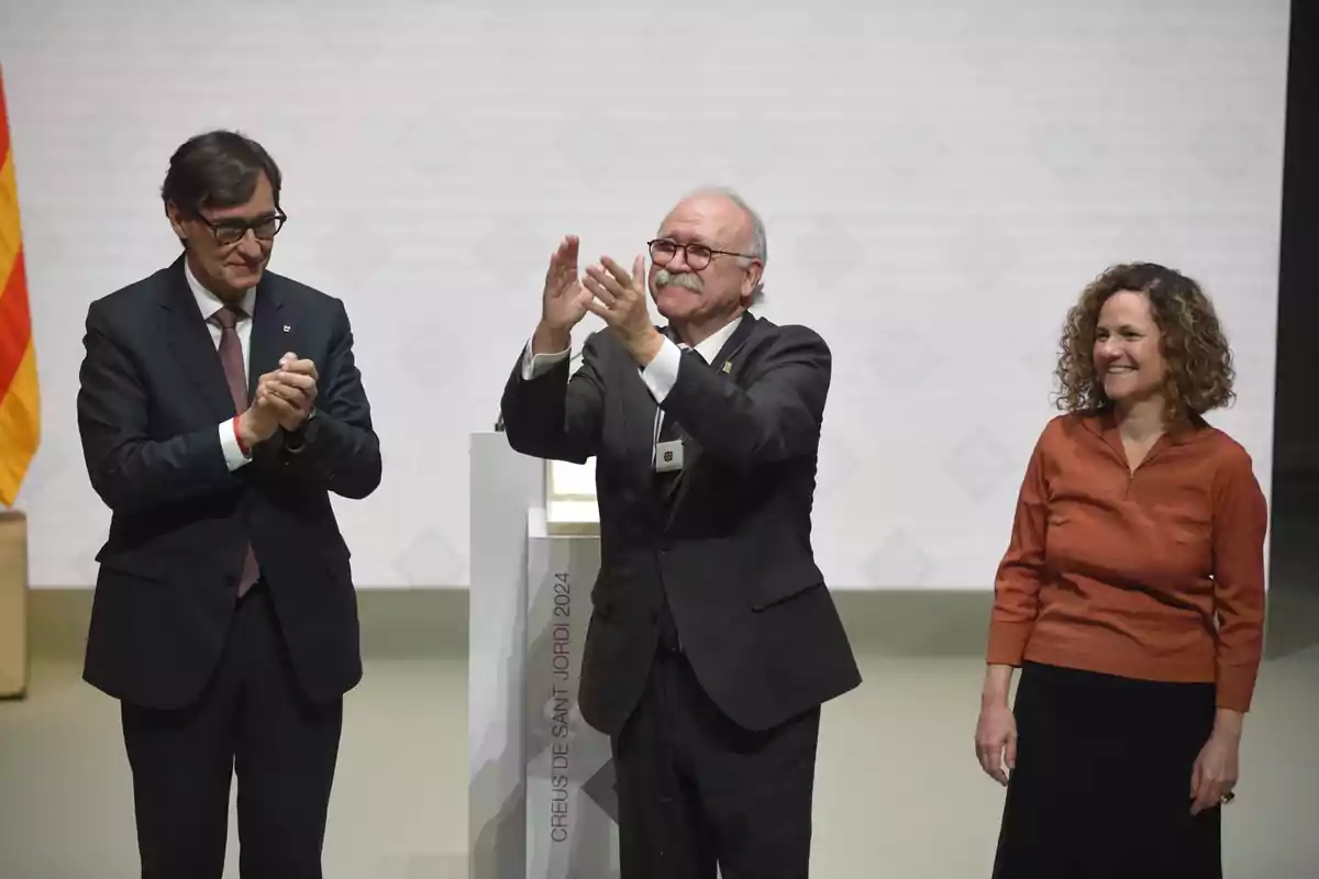 Tres personas de pie en un escenario durante una ceremonia, una de ellas aplaudiendo, otra levantando las manos y la tercera sonriendo, con una bandera catalana al fondo y un pedestal con la inscripción Creus de Sant Jordi 2024. Tres personas de pie en un escenario durante una ceremonia, una de ellas aplaudiendo, otra levantando las manos y la tercera sonriendo, con una bandera catalana al fondo y un pedestal con la inscripción Creus de Sant Jordi 2024.