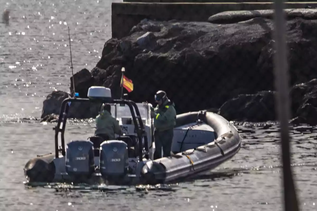 Una lancha neumática con dos motores y bandera de España navega cerca de unas rocas con dos personas a bordo vestidas con uniformes verdes Una lancha neumática con dos motores y bandera de España navega cerca de unas rocas con dos personas a bordo vestidas con uniformes verdes