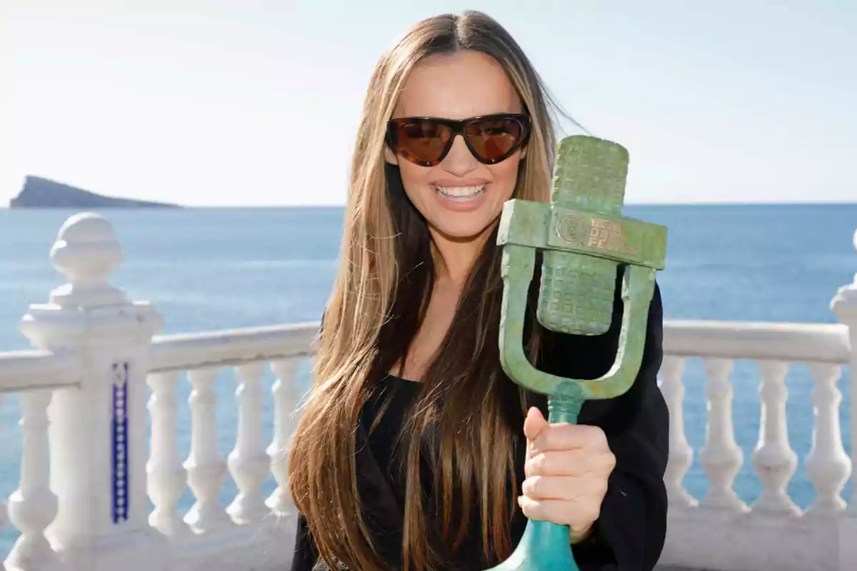 Mujer sonriente con gafas de sol sostiene un trofeo en forma de micrófono frente al mar y una barandilla blanca Mujer sonriente con gafas de sol sostiene un trofeo en forma de micrófono frente al mar y una barandilla blanca