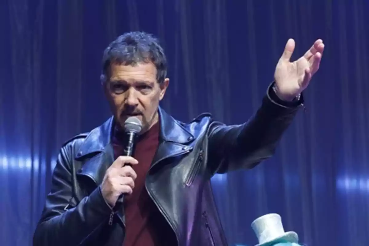 Hombre con chaqueta de cuero negro hablando por micrófono y levantando una mano en un escenario iluminado con fondo azul Hombre con chaqueta de cuero negro hablando por micrófono y levantando una mano en un escenario iluminado con fondo azul