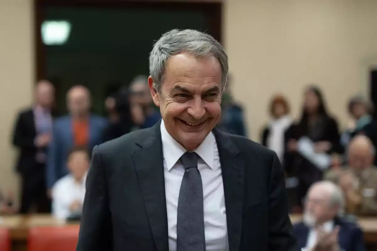Hombre de cabello canoso con traje oscuro y corbata gris sonriendo en un evento con personas desenfocadas al fondo Hombre de cabello canoso con traje oscuro y corbata gris sonriendo en un evento con personas desenfocadas al fondo