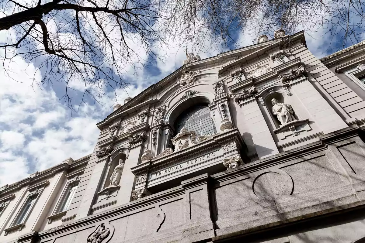 Fachada del Tribunal Supremo de España vista desde abajo con esculturas y ramas de árboles enmarcando el edificio bajo un cielo parcialmente nublado Fachada del Tribunal Supremo de España vista desde abajo con esculturas y ramas de árboles enmarcando el edificio bajo un cielo parcialmente nublado