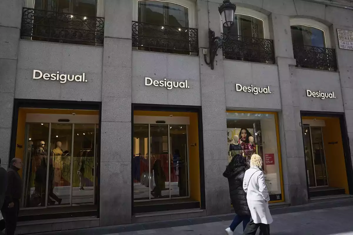 Fachada de una tienda Desigual con varias personas caminando por la acera en un día soleado Fachada de una tienda Desigual con varias personas caminando por la acera en un día soleado
