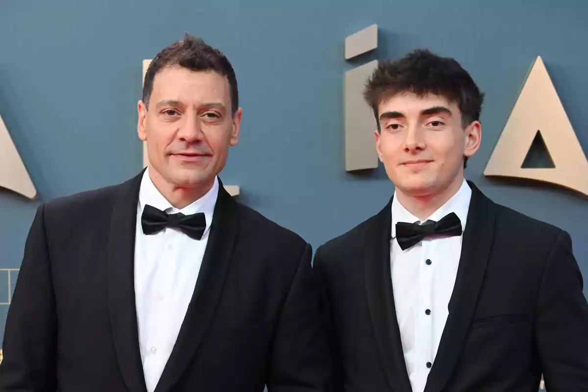 Dos hombres vestidos con esmoquin negro y pajarita posan juntos en una alfombra roja frente a un fondo azul con letras doradas