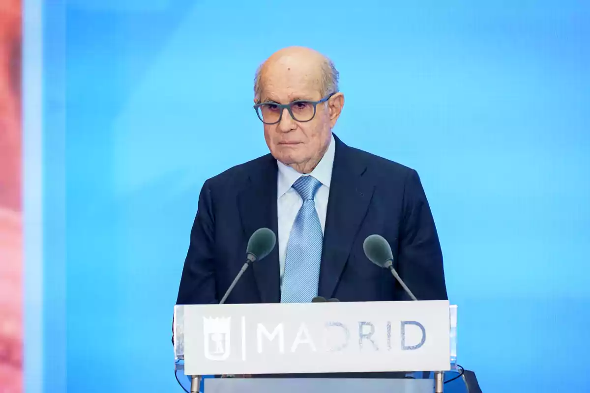 Hombre mayor con gafas y traje oscuro hablando en un atril con la palabra Madrid sobre fondo azul Hombre mayor con gafas y traje oscuro hablando en un atril con la palabra Madrid sobre fondo azul