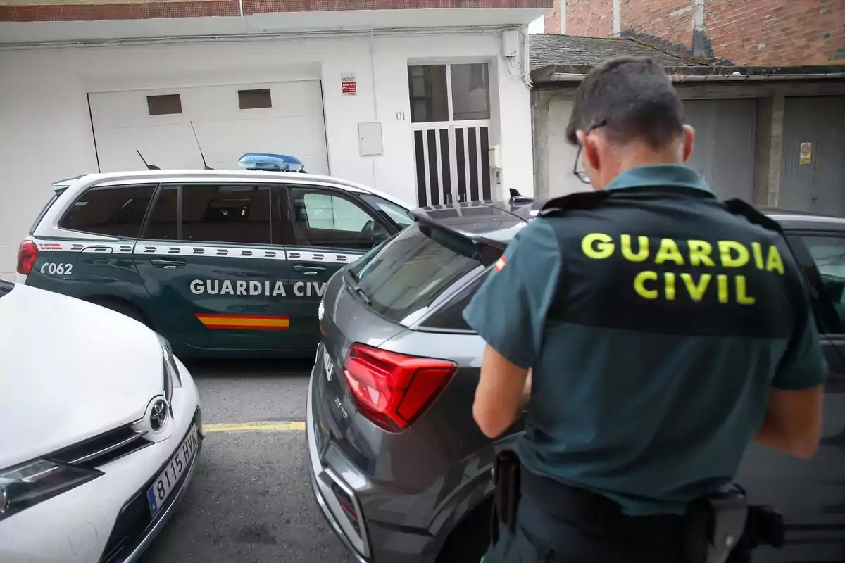Un agente de la Guardia Civil de espaldas junto a varios coches estacionados en una calle frente a un edificio Un agente de la Guardia Civil de espaldas junto a varios coches estacionados en una calle frente a un edificio