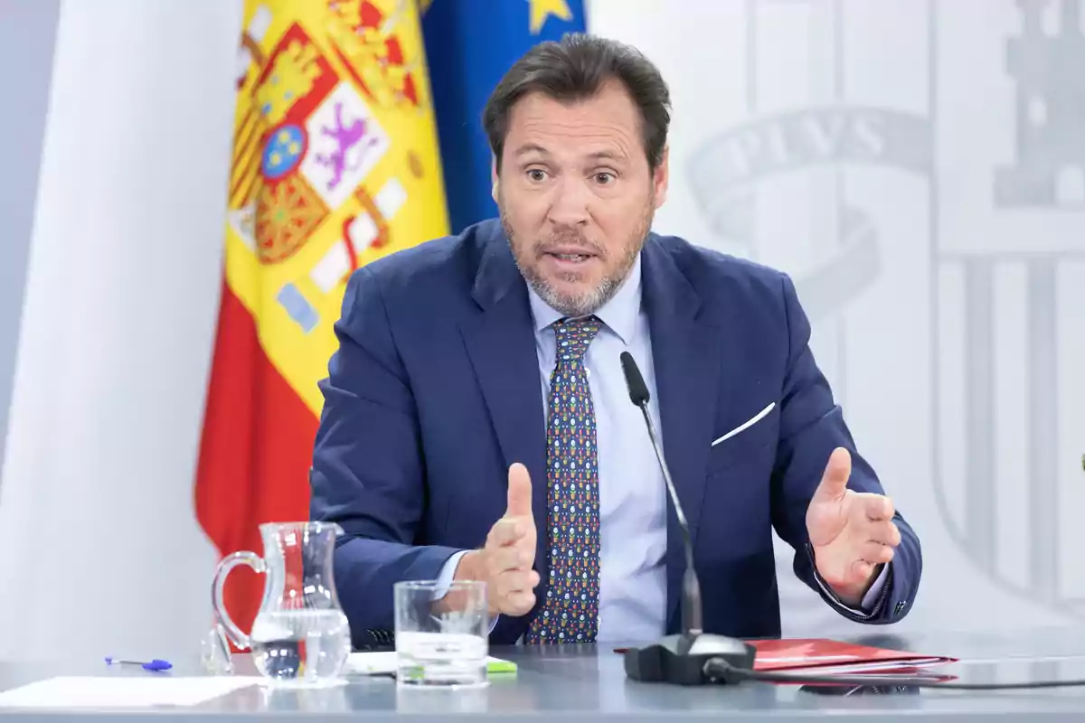Hombre con traje azul hablando en una conferencia de prensa con una bandera de España al fondo