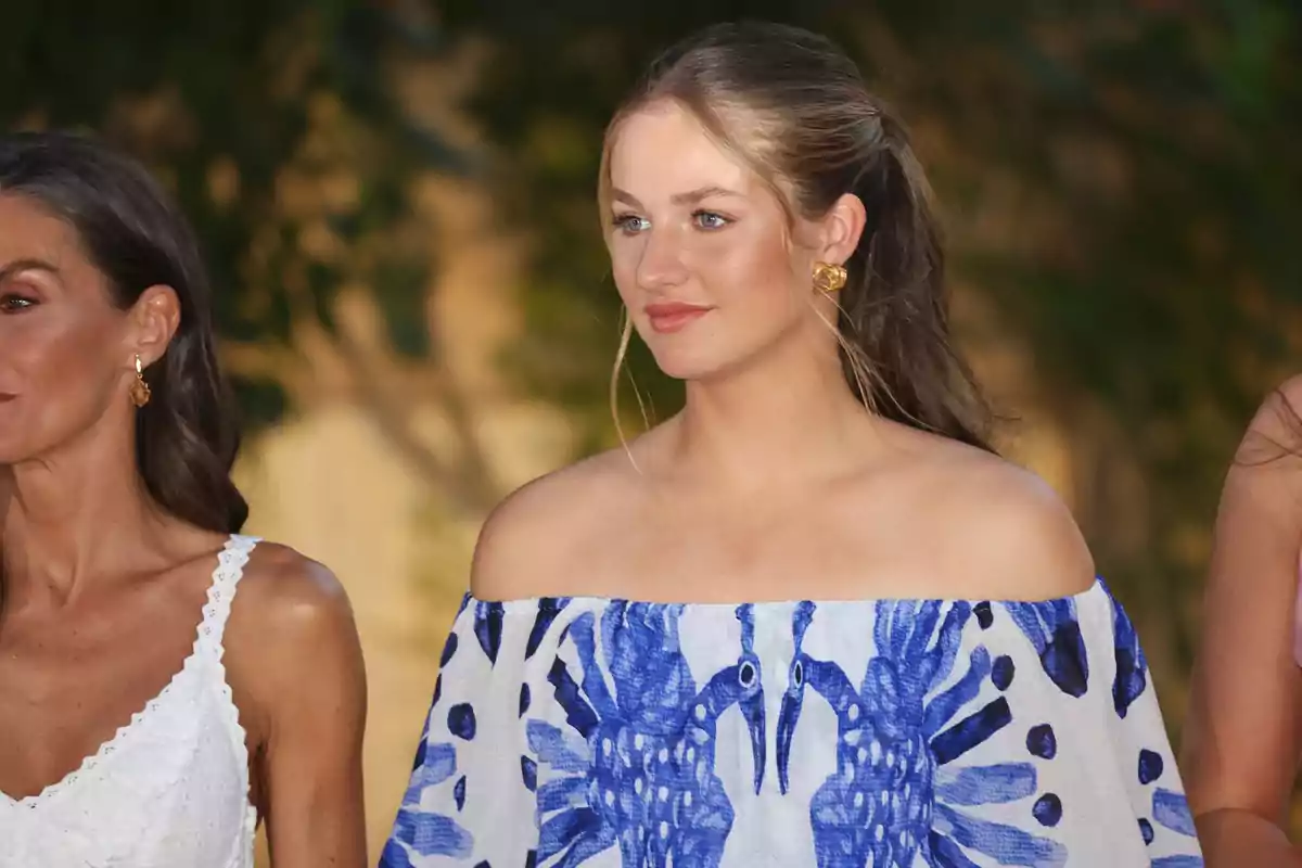 Dos mujeres posan al aire libre, una de ellas lleva un vestido blanco y la otra un vestido azul y blanco con hombros descubiertos y estampado de aves Dos mujeres posan al aire libre, una de ellas lleva un vestido blanco y la otra un vestido azul y blanco con hombros descubiertos y estampado de aves