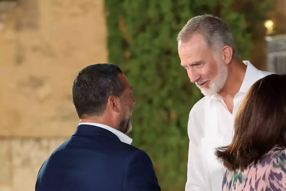 Dos hombres conversan sonrientes en un evento al aire libre mientras una mujer de espaldas aparece en primer plano Dos hombres conversan sonrientes en un evento al aire libre mientras una mujer de espaldas aparece en primer plano