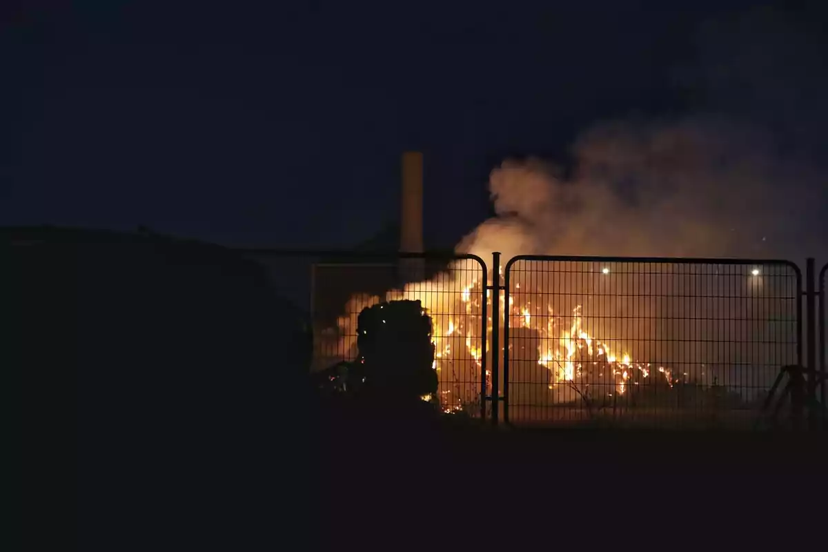 Fuego ardiendo detrás de una cerca metálica durante la noche con humo elevándose al cielo oscuro Fuego ardiendo detrás de una cerca metálica durante la noche con humo elevándose al cielo oscuro