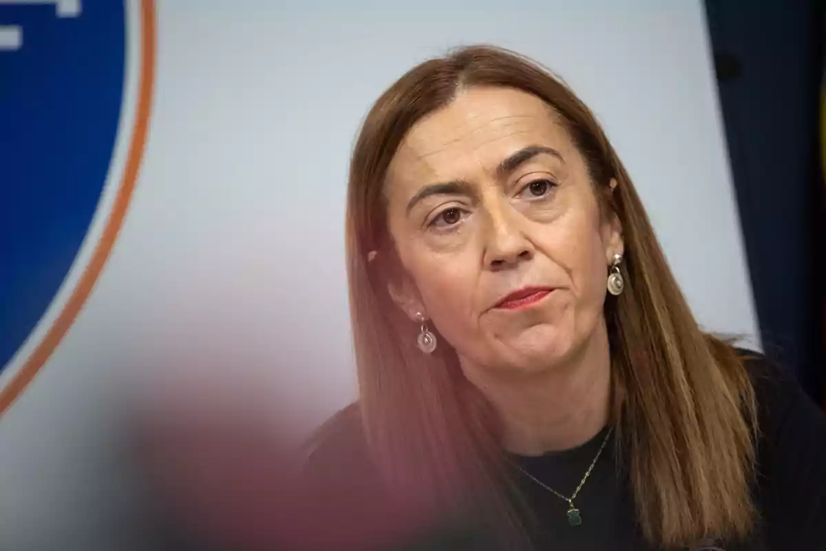 Mujer de cabello lacio castaño claro con expresión seria usando pendientes y collar sentada frente a un fondo blanco y azul