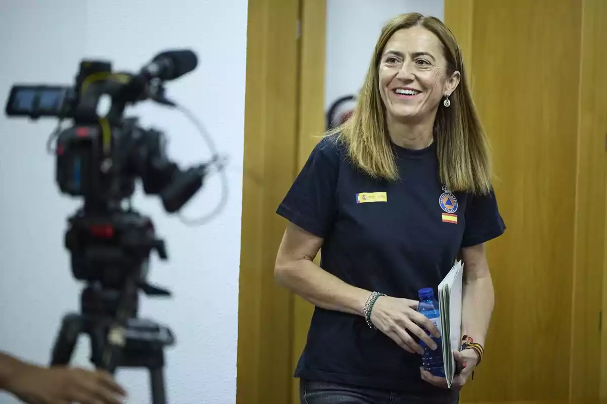 Mujer sonriente con camiseta de protección civil sosteniendo una botella de agua y unos documentos mientras una cámara de video la graba en un interior Mujer sonriente con camiseta de protección civil sosteniendo una botella de agua y unos documentos mientras una cámara de video la graba en un interior