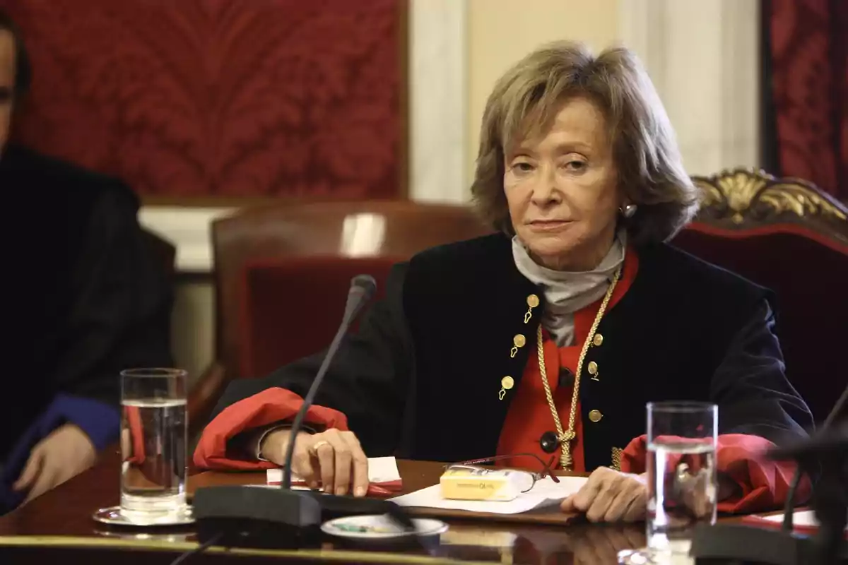 Mujer mayor con toga judicial sentada en una mesa de madera con micrófono y vasos de agua en una sala elegante Mujer mayor con toga judicial sentada en una mesa de madera con micrófono y vasos de agua en una sala elegante